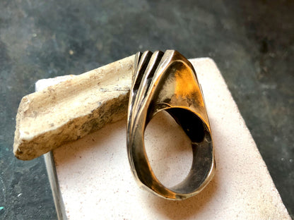 Anello materico in bronzo con un design ondulato, posizionato su una superficie di pietra chiara