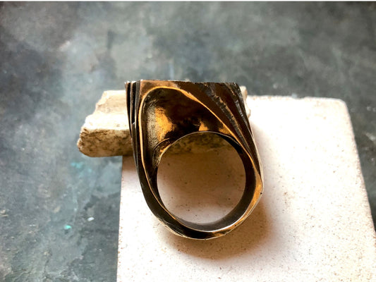 Anello materico in bronzo con design ondulato, posato su una superficie neutra