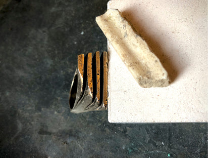 Anello materico in bronzo con dettagli strutturali vicino a una pietra chiara su sfondo scuro