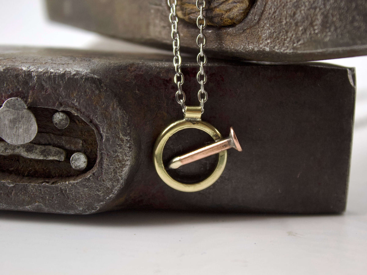 Collana chiodo con pendente a cerchio in ottone e chiodo in rame, appoggiata su un supporto scuro