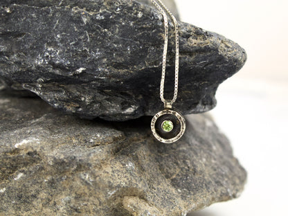 Collana in argento con ciondolo circolare che mostra un peridoto verde al centro, su una roccia scura
