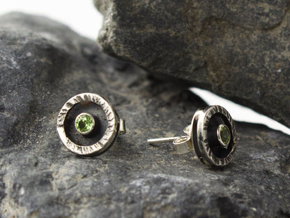 Orecchini con peridoto di 3mm in cornice circolare con superficie martellata, posizionati su una superficie rocciosa.