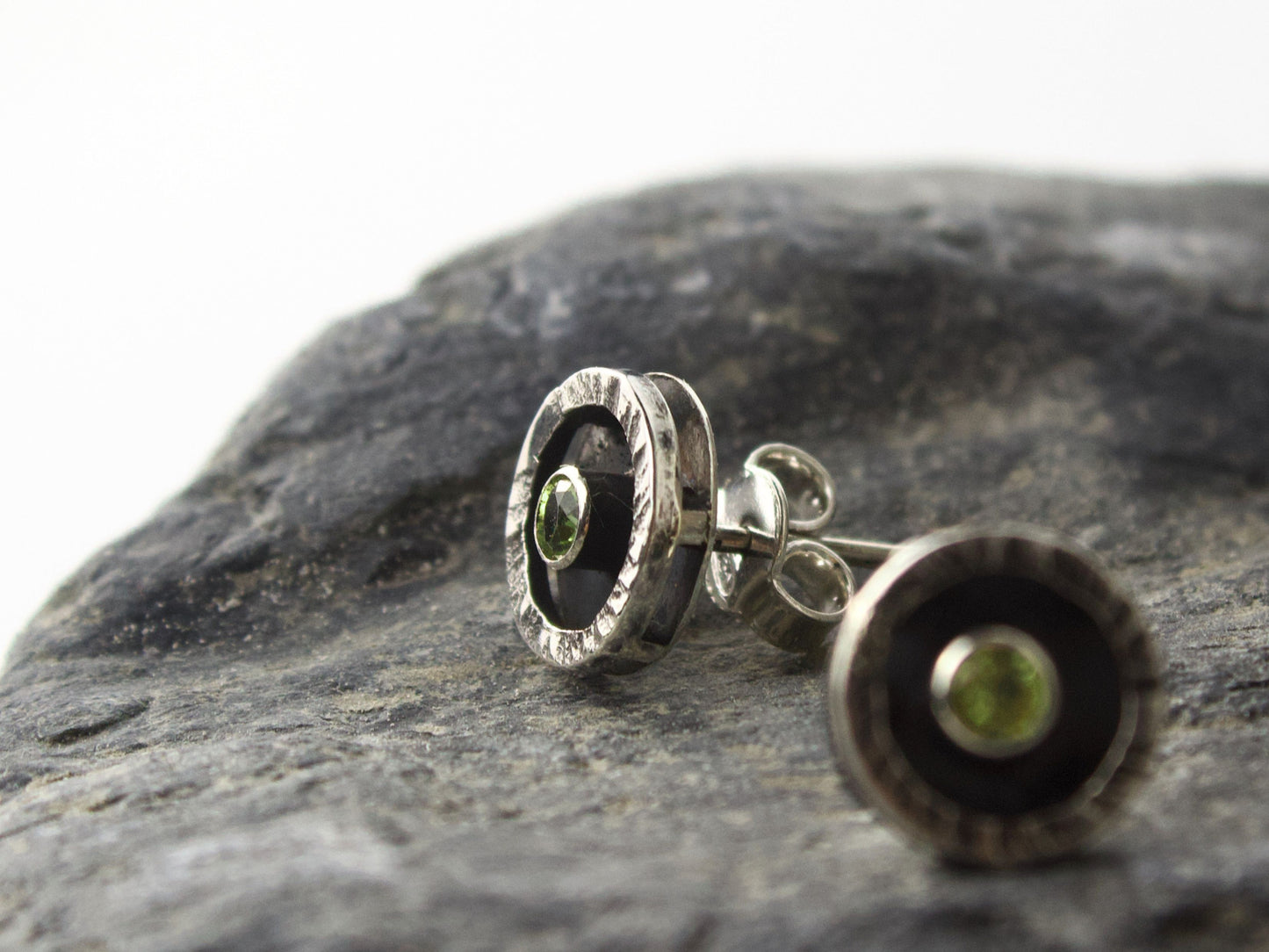 Orecchini Glimoire con peridoto da 3mm Ø, cornice circolare martellata in argento, sopra una pietra scura.