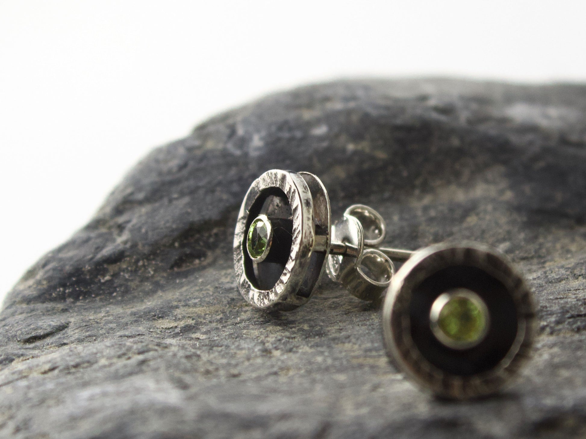 Orecchini Glimoire con peridoto da 3mm Ø, cornice circolare martellata in argento, sopra una pietra scura.