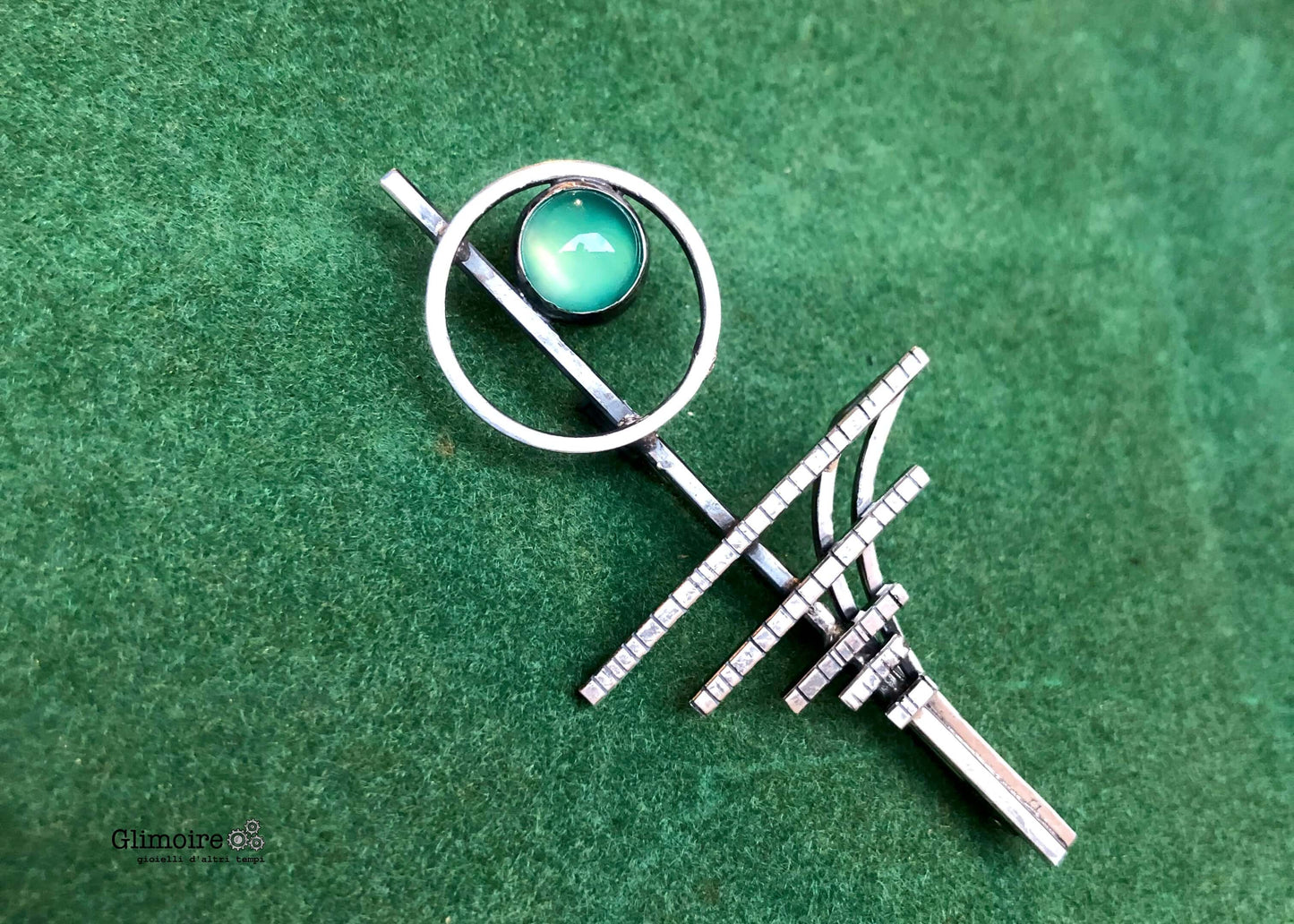 Spilla elegante con pietra verde su sfondo verde, con dettagli geometrici e finiture lucide
