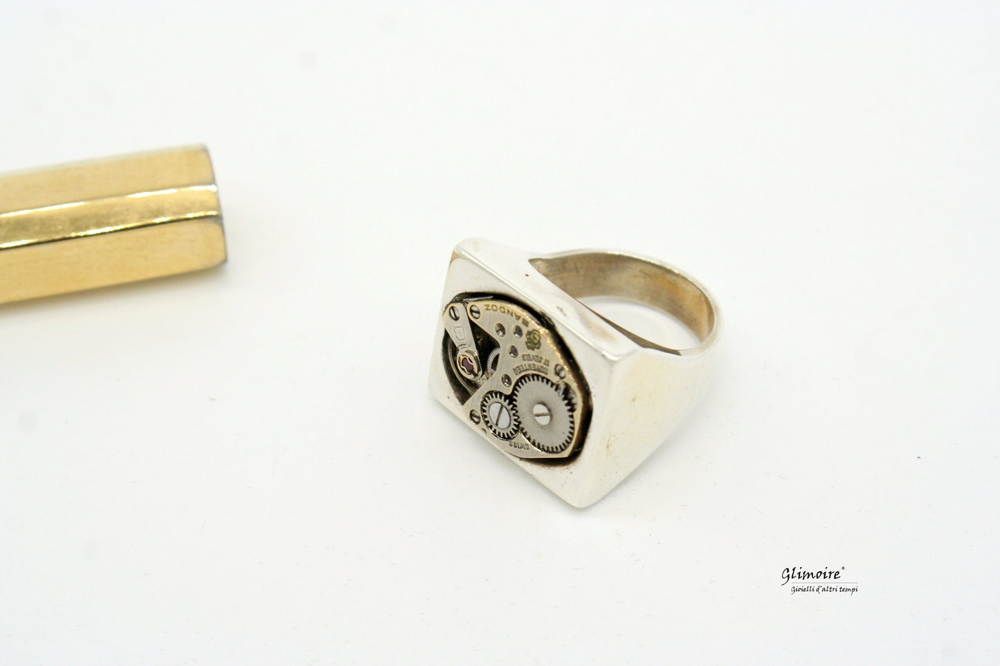 Anello in argento con meccanismo dettagliato e design unico, accanto a un oggetto dorato